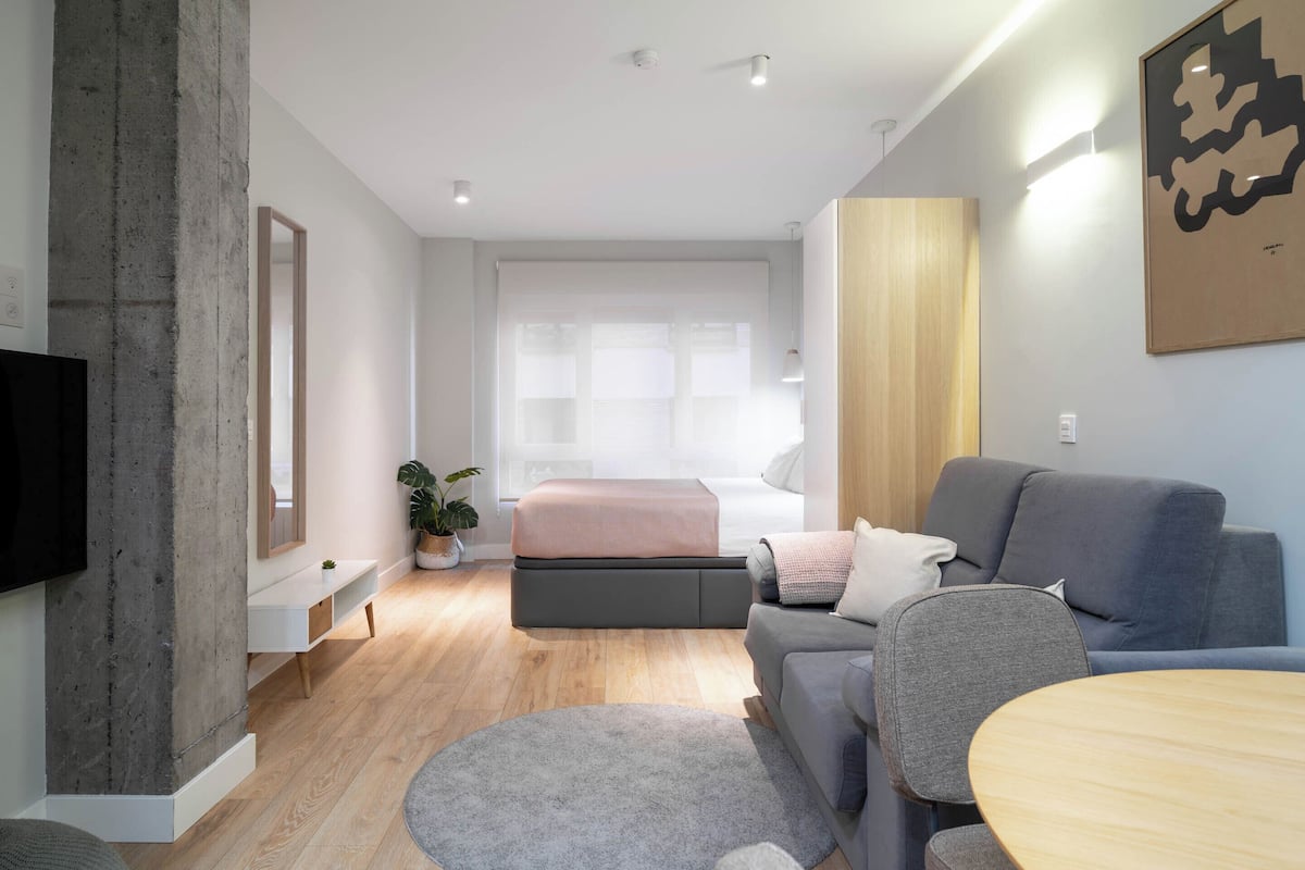 Una sala de estar moderna con un sofá gris, una mesa redonda de madera, un tapete gris, un ventanal con cortinas translúcidas y un televisor de pantalla plana montado en la pared.