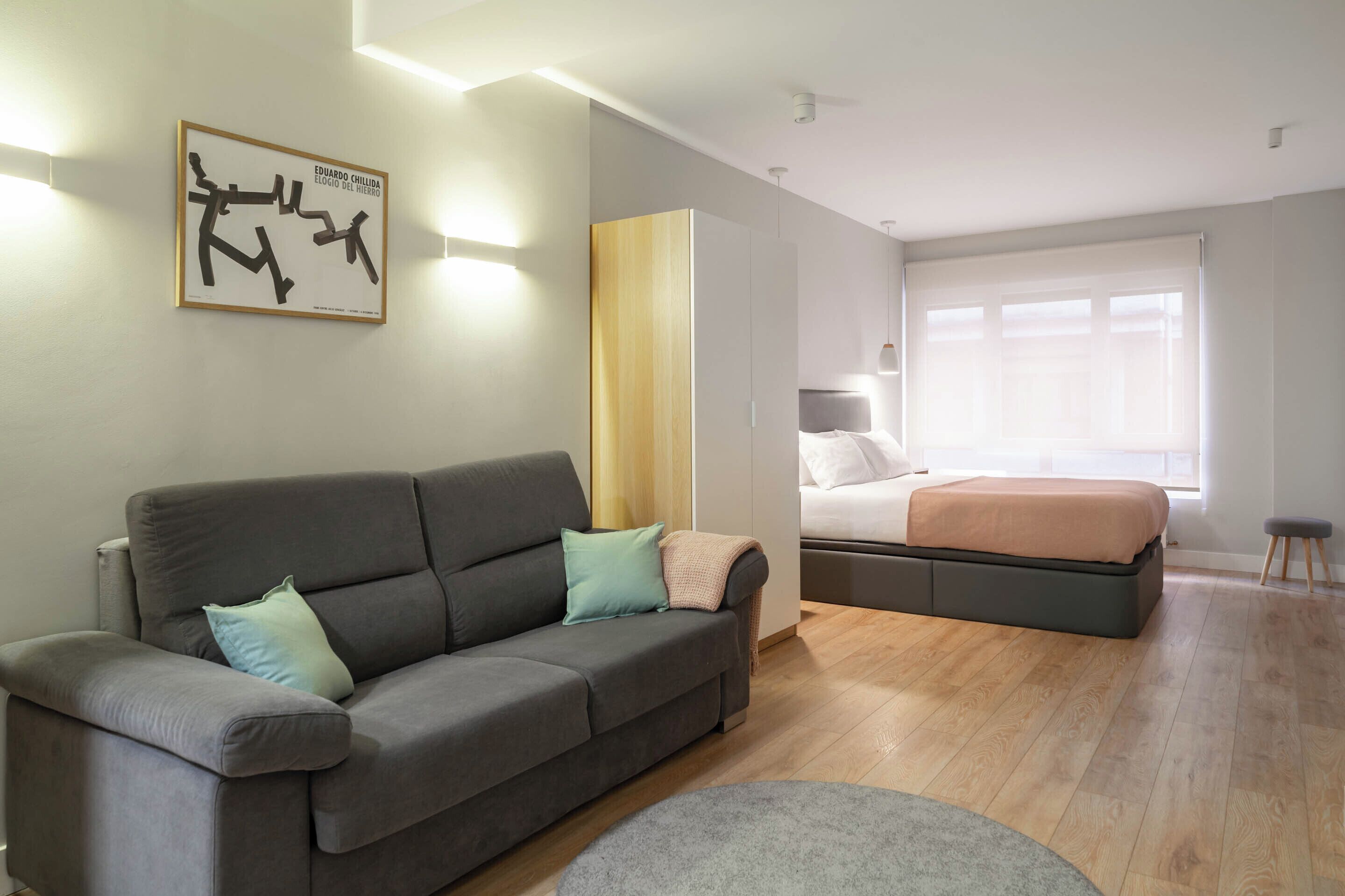 A modern bedroom with a grey sofa, a bed, and a large window.