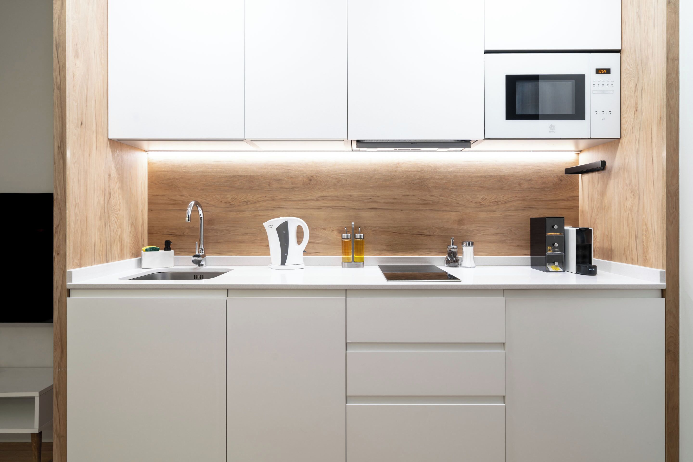 Una cocina moderna con armarios blancos, microondas integrado, hervidor de agua y cafetera.