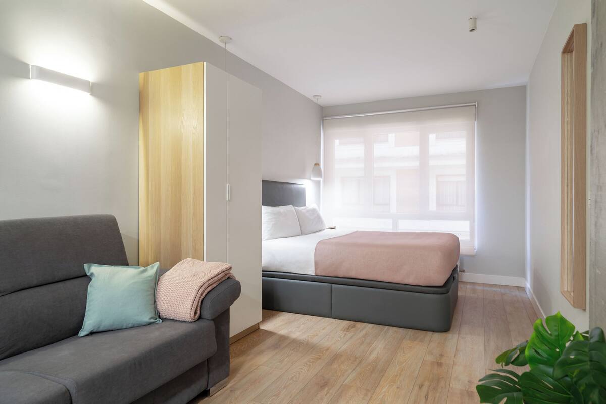 Una habitación moderna y bien iluminada con un sofá gris, una cama con cabecero gris y un armario de madera.
