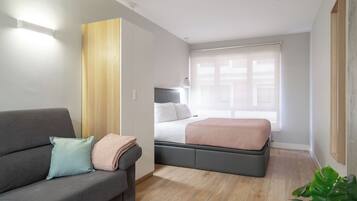 Una habitación moderna y bien iluminada con un sofá gris, una cama con cabecero gris y un armario de madera.