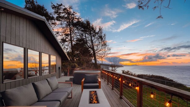 A deck with a sofa, a coffee table, and a fire pit, overlooking a body of water with a sunset view. - Private Blufftop · Oceanview Hot Tub · Fire Pit (Port Angeles)