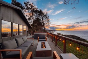 A deck with a sofa, a coffee table, and a fire pit, overlooking a body of water with a sunset view.