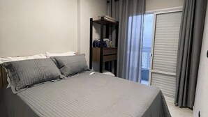 A neatly made bed with a grey bedspread, a bedside table with a lamp, and a window with grey curtains. - UNITA 157 (Praia Grande)