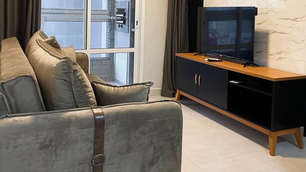 A modern living room with a grey sofa, a black entertainment unit, and a large window with curtains. - UNITA 157 (Praia Grande)