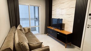 A modern living room with a beige sofa, a flat-screen TV on a wooden cabinet, and a large window with curtains. - UNITA 157 (Praia Grande)