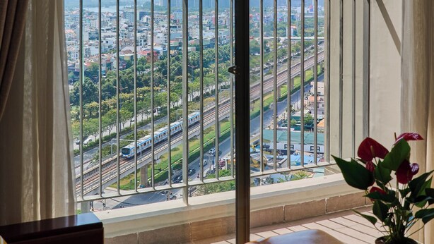 A balcony with a cityscape view, including a prominent skyscraper, a train, and a well-maintained garden area. - Masteri view Landmark 81, downtown city 5mins-metro (Hồ Chí Minh)
