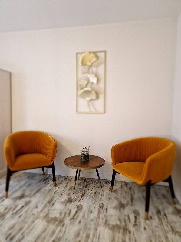 Two orange armchairs, a small round table with a candle holder, and a wall-mounted abstract art piece. - Une autre maison (Pupillin)