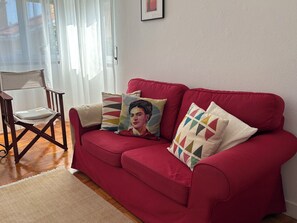 Un sofá rojo con cojines estampados, una silla de madera y una alfombra beige en una habitación con pared blanca y una ventana con cortinas de encaje.