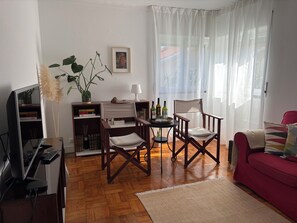 Una sala de estar con piso de madera, un sofá rojo, un televisor, una estantería, una mesa de comedor con sillas y una ventana con cortinas traslúcidas.