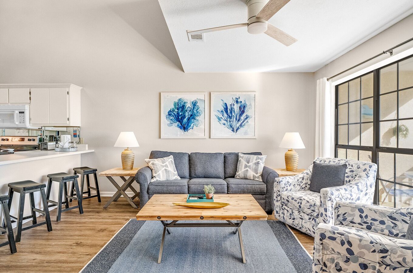 Een moderne woonkamer met een grijze bank, een houten salontafel en twee fauteuils met een patroon. Er is een keukenhoek met witte kastjes en een magnetron. Twee abstracte blauwe schilderijen hangen aan de muur.