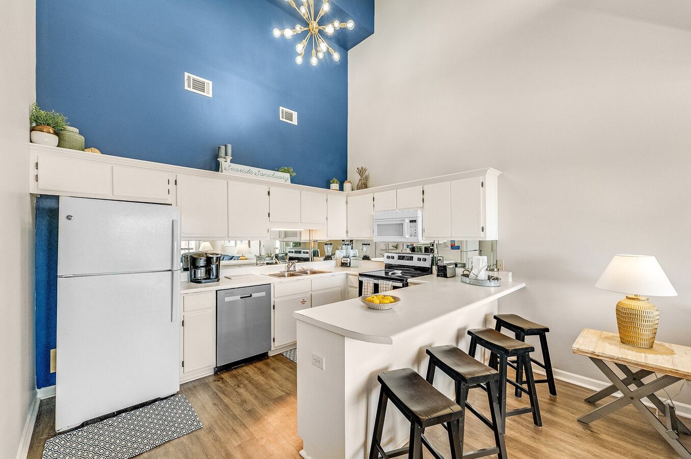Cozinha moderna com armários brancos, uma parede de destaque azul, uma ilha central com banquetas e um lustre.