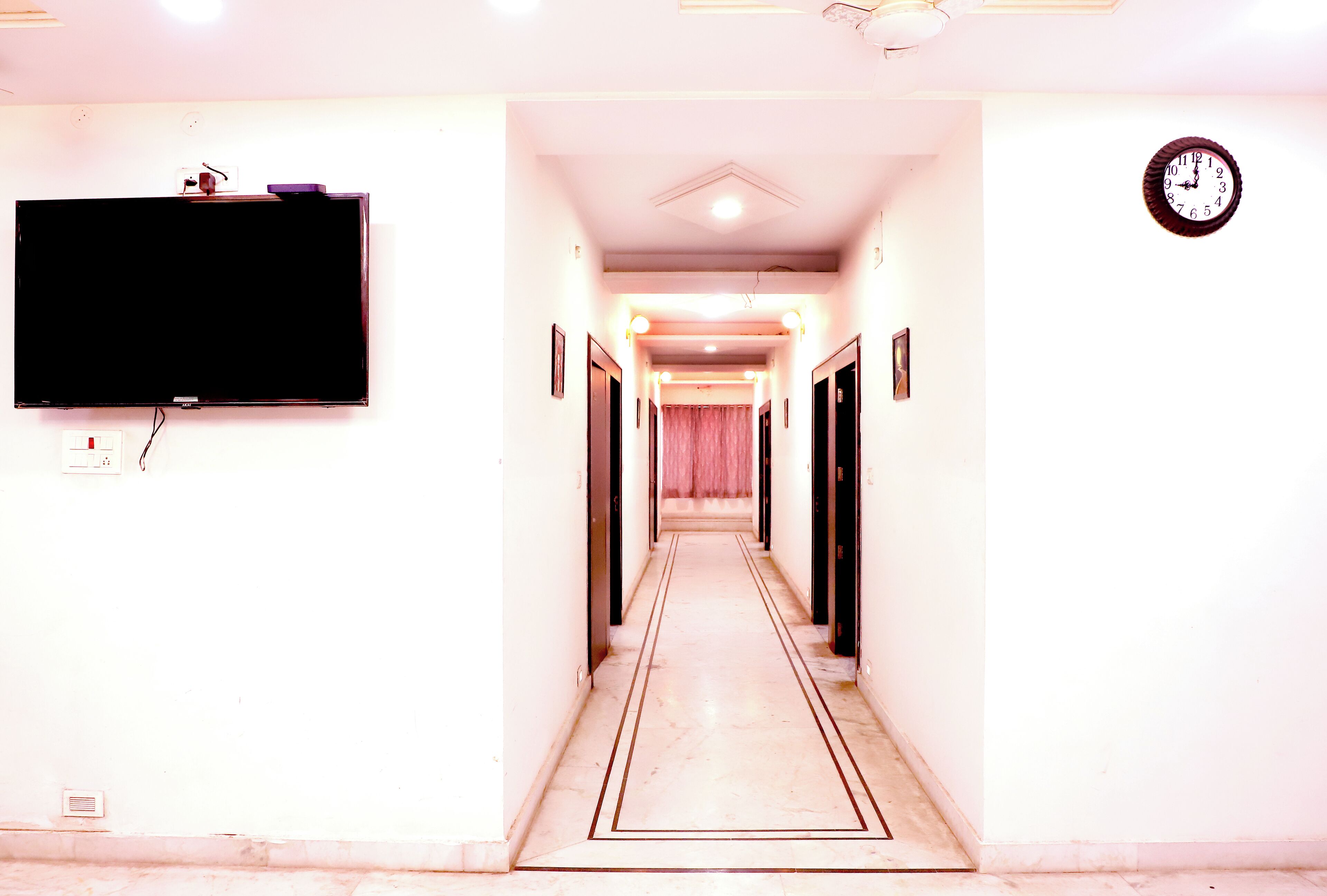 A hotel hallway with a flat-screen TV mounted on the wall, a clock on the right, and a door at the end.