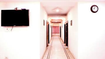 A hotel hallway with a flat-screen TV mounted on the wall, a clock on the right, and a door at the end.