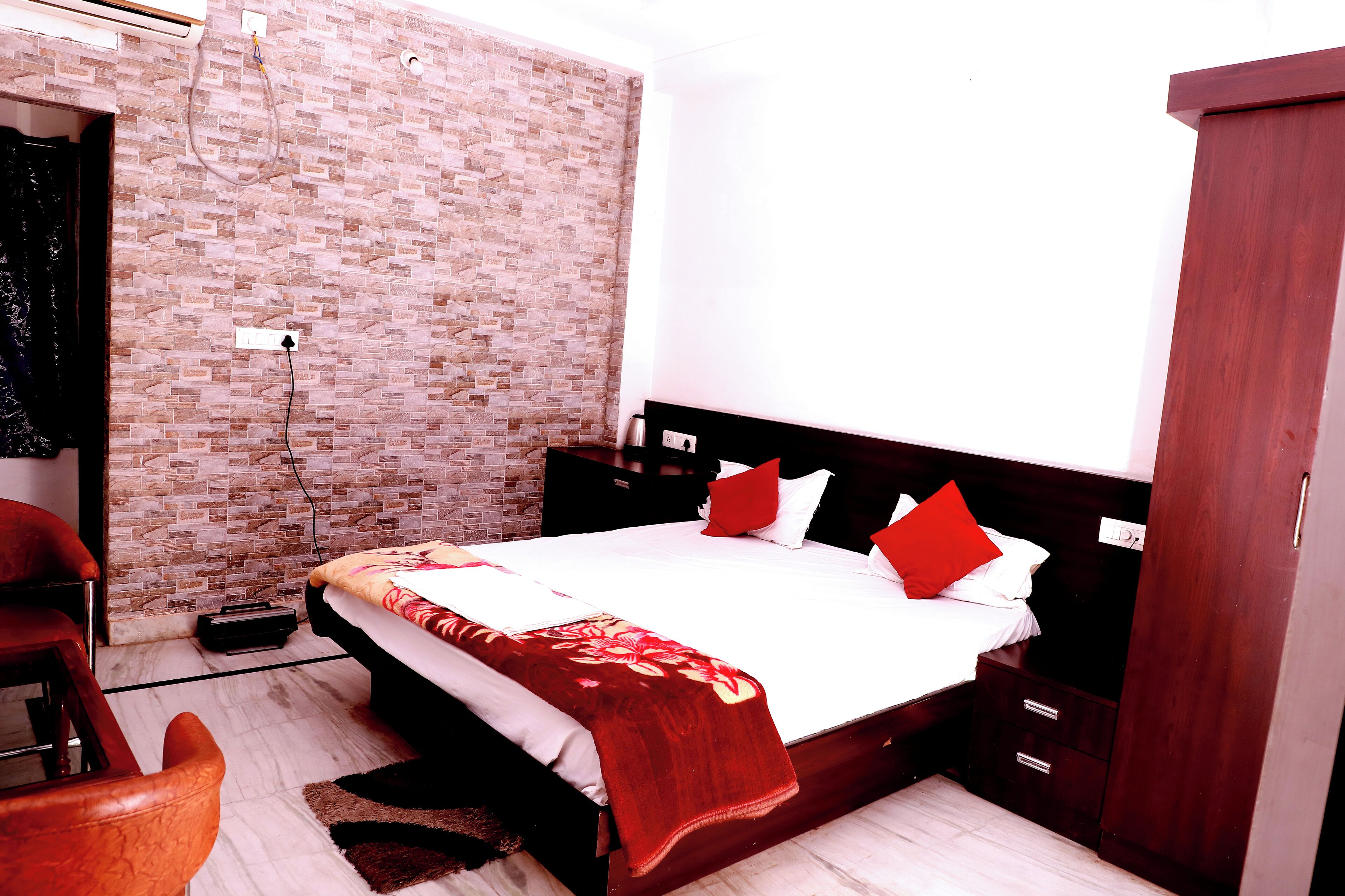 A bedroom with a brick-patterned wall, a bed with white bedding and red pillows, a wooden nightstand, and a red throw blanket.