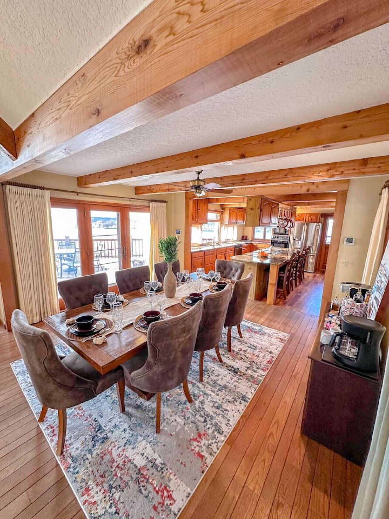 A spacious dining area with a wooden table set for six, surrounded by upholstered chairs. A kitchen is visible in the background with wooden cabinets and a central island.