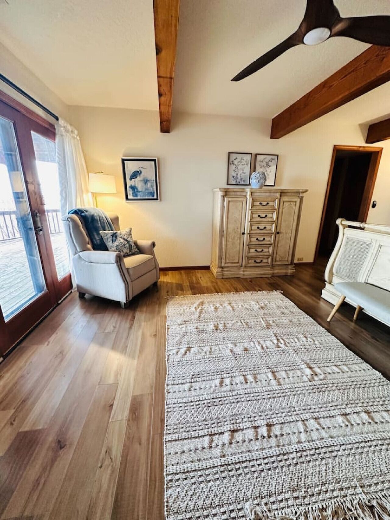 A cozy living room with a wooden floor, a white sofa, a chair, a large rug, and a cabinet with artwork.