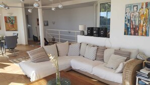 Un salon spacieux avec un grand canapé blanc, une table basse en bois et un vase rempli de fleurs. On y trouve également un coin repas avec des chaises et une table, ainsi qu’un tableau accroché au mur.