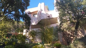 Un bâtiment à la façade blanche, entouré d’un jardin où poussent différentes plantes et arbres.