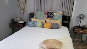 Una cama bien hecha con una colcha a rayas y una cesta tejida, un atrapasueños colgado en la pared, y una variedad de almohadas estampadas.