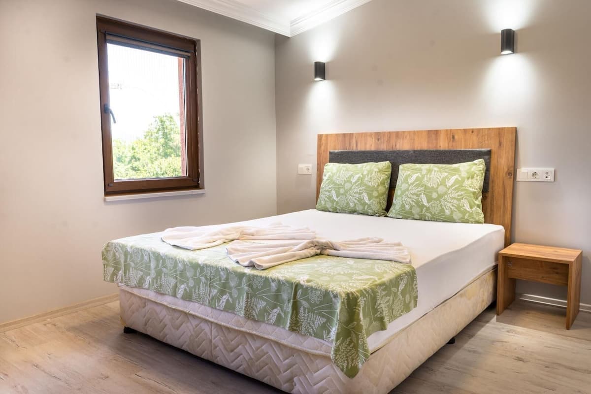 Una cama bien hecha con una colcha floral verde, un cabecero de madera y dos almohadas verdes. Junto a la cama hay una mesita de noche de madera.