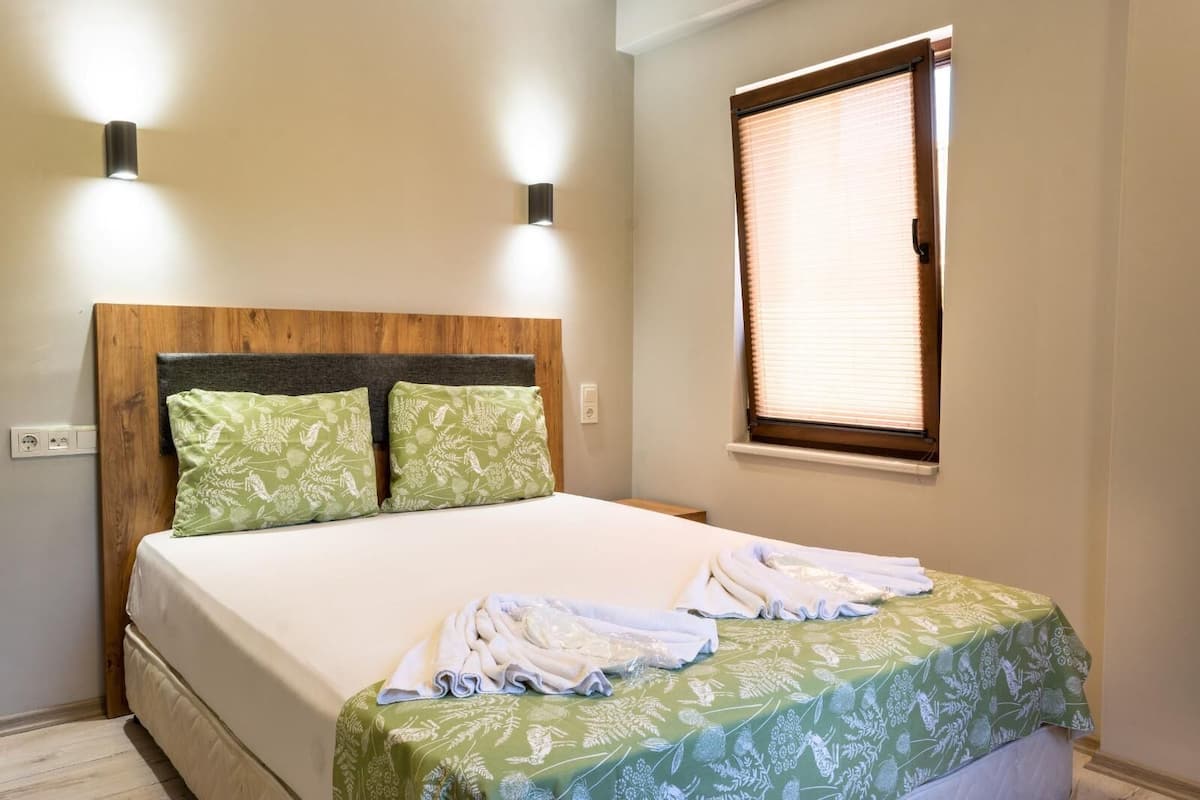 Una cama bien hecha con una colcha floral verde, dos almohadas a juego y un cabecero de madera. A la derecha de la cama, hay una ventana con persianas.
