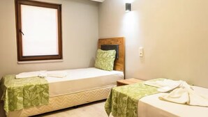 Une petite chambre propre, avec deux lits simples, chacun recouvert d’une couverture verte et agrémenté d’oreillers blancs. On y trouve une fenêtre avec des stores, une table de chevet et une lampe fixée au mur.