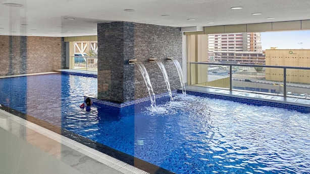 Ein Innenpool mit mehreren Wasserstrahlen, einem gefliesten Boden und Blick auf die Stadt durch große Fenster.