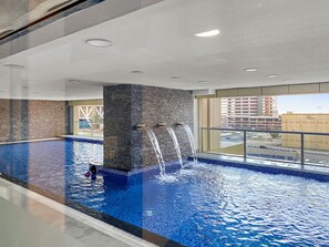 An indoor swimming pool with multiple water jets, a tiled floor, and a view of the city through large windows. - Stylish 1BR with Balcony | Near Marina Mall, Pool & Free Parking (Marina)