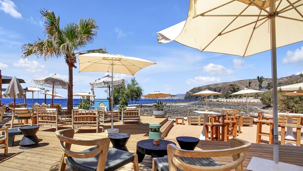 Eine Holzterrasse mit Sitzgelegenheiten im Freien, Sonnenschirmen und Blick auf Strand und Meer.