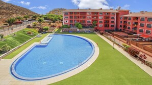 Ein Schwimmbecken mit umlaufender Rasenfläche, ein rotes Gebäude mit Balkonen und eine hügelige Landschaft im Hintergrund.