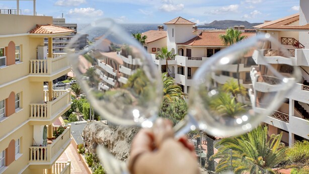 A view through a glass window of a residential area with balconies, a rocky outcrop, and a distant mountain range. - Sun And Green, Heated Pool, 3 Rooms, Air C (San Miguel de Abona)