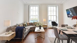 Una sala de estar moderna con un sofá modular, una mesa de comedor con sillas, un televisor de pantalla plana montado en la pared y ventanas amplias que dejan entrar luz natural.