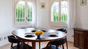 Une salle à manger avec une table ronde, des chaises noires et un vase rempli de fleurs jaunes. Deux fenêtres, ornées de rideaux, sont visibles, et un buffet se trouve sur le côté droit.