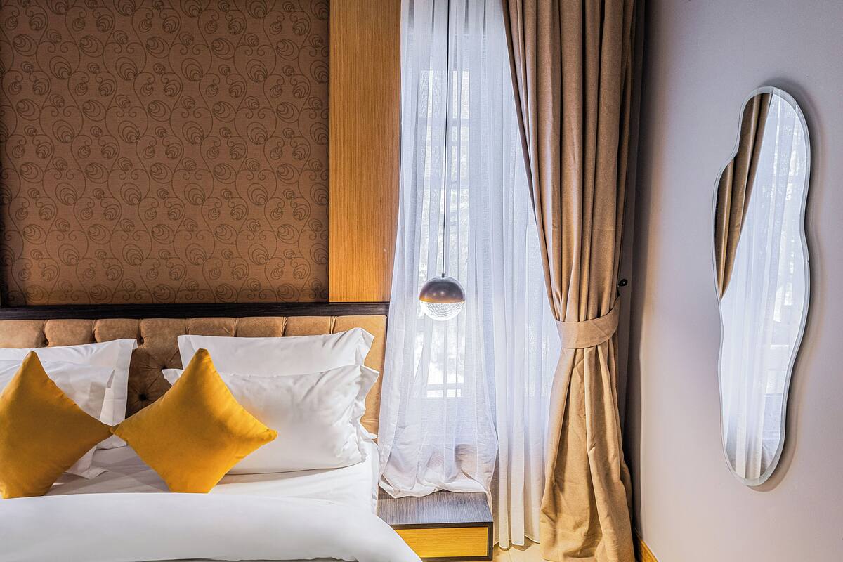 A hotel room with a tufted headboard, a bed with white and mustard pillows, sheer curtains, and a wall-mounted mirror.