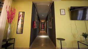 A hotel corridor with a long, narrow hallway, a fire extinguisher, a framed artwork, a television mounted on the wall, and a small table with a vase. - amp lodge (Kumasi)