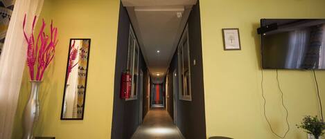 A hotel corridor with a long, narrow hallway, a fire extinguisher, a framed artwork, a television mounted on the wall, and a small table with a vase.