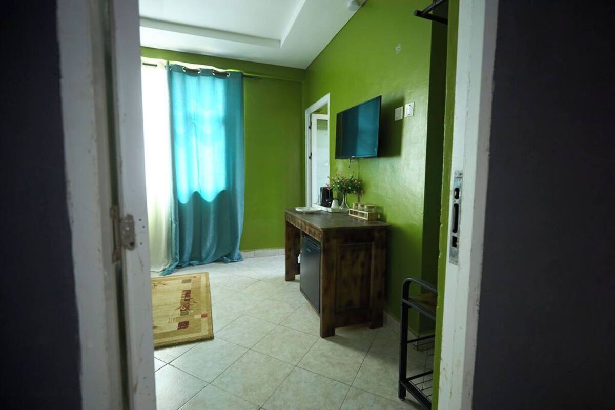 Una habitación con pared verde, un escritorio de madera, una silla, un televisor fijado a la pared y una cortina azul.