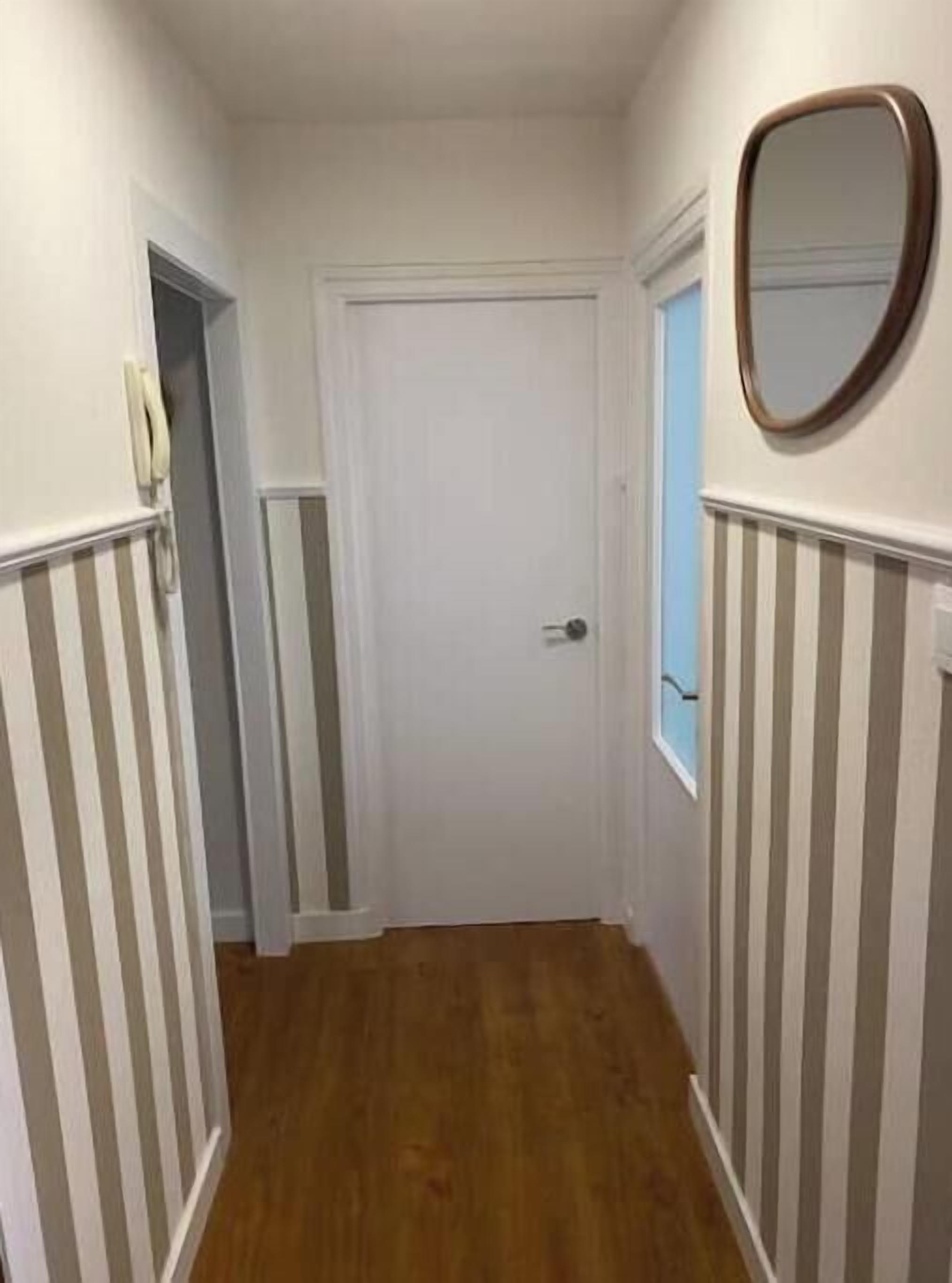 Un couloir étroit aux murs recouverts d’un papier peint à rayures, une porte blanche et un miroir accroché au mur.