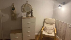 Een kamer met een houten vloer, een beige fauteuil met een kussen, een witte dressoir met lades, een spiegel aan de wand en een wandlamp.