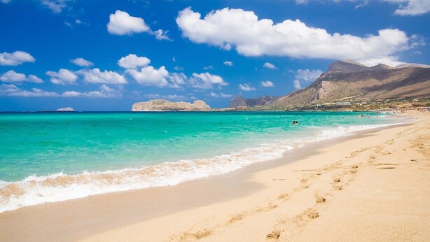 A beach with clear turquoise water, sandy shore, and rocky mountains in the background.