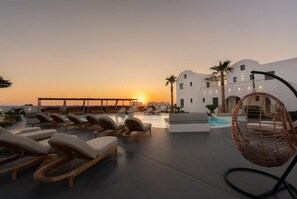 A rooftop pool area with lounge chairs and a hanging chair, overlooking a sunset. - Amethyst Luxury Suites - Oia Junior Suite (Santorini)