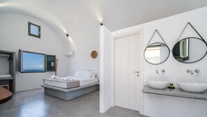 A modern bedroom with a bed, a large window, and a bathroom with two sinks and mirrors. - Amethyst Luxury Suites - Oia Junior Suite (Santorini)