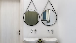 A bathroom with two white sinks, a door, and two round mirrors mounted on the wall. - Amethyst Luxury Suites - Oia Junior Suite (Santorini)