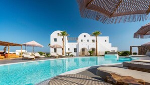 A pool area with lounge chairs and umbrellas, a white building with arches, and a clear blue sky. - Amethyst Luxury Suites - Oia Junior Suite (Santorini)