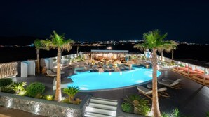 A nighttime view of a rooftop pool area with lounge chairs, palm trees, and a bar area.