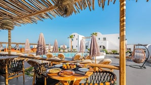 A rooftop dining area with tables and chairs, a pool, and white buildings in the background.