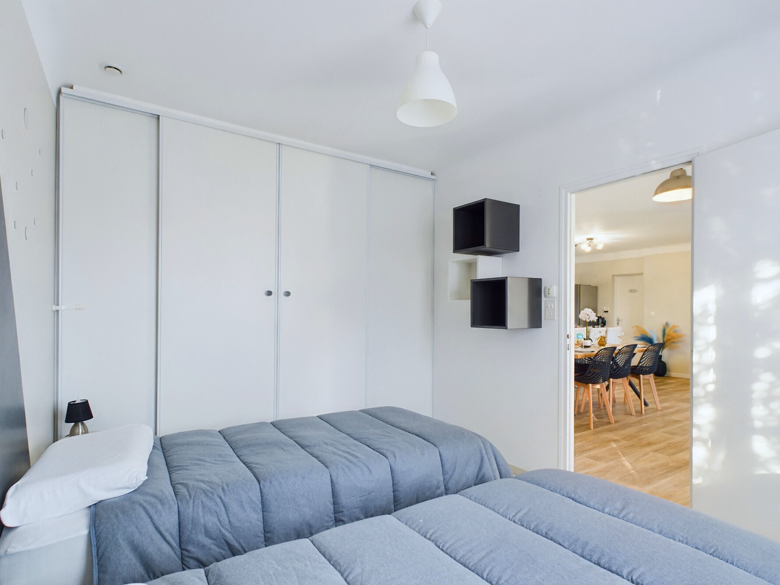 A bedroom with a bed, a wardrobe, and a dining area visible through an open door.