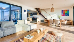 A modern living room with a sofa, coffee table, and dining area with chairs. There is a staircase leading to an upper level. - Pool, view and beach access, 6 adults/2 children (La Guérinière)