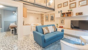 A modern living room with a blue sofa, a dining area, and a TV. - Cinque Giornate 70 Suites by Rent all Como (Como)